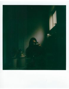 A polaroid of a person with long hair, taking a photo of themselves in the mirror of a shadowy bathroom.