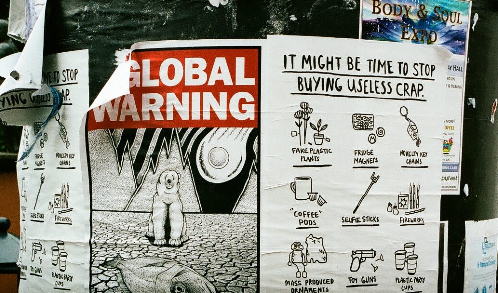 Photo of posters stuck to a pillar. One has a black and white illustration of a polar bear and a dead fish in a dry desert landscape with a flaming sun behind them, with the words "global warming" at the top against a red background. Next to it is another poster that says "it might be time to stop buying useless crap", with illustrations of fake plastic plants, fridge magnets, novelty key chains, "coffee" pods, selfie sticks, fireworks, mass produced ornaments, toy guns and plastic party cups.