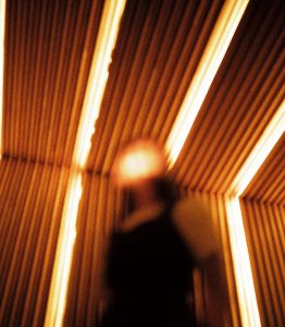 Blurred photo of a person with long hair from the waist up, taken from below. Their face is blurred out and lit up so the features aren't visible. The background is a wall and roof with some parts lit up an orangey colour.